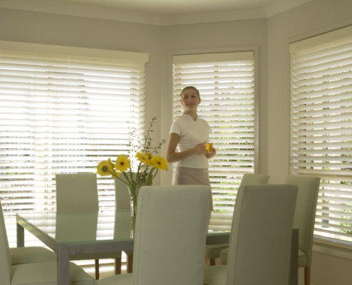 Woman holding glass of water standing behind the kitchen table appreciating the light control done by Timberstyle venetian blinds