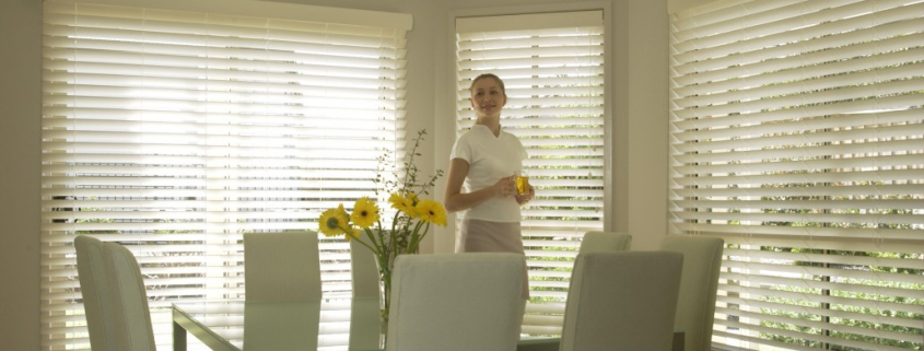 Woman holding glass of water standing behind the kitchen table appreciating the light control done by Timberstyle venetian blinds